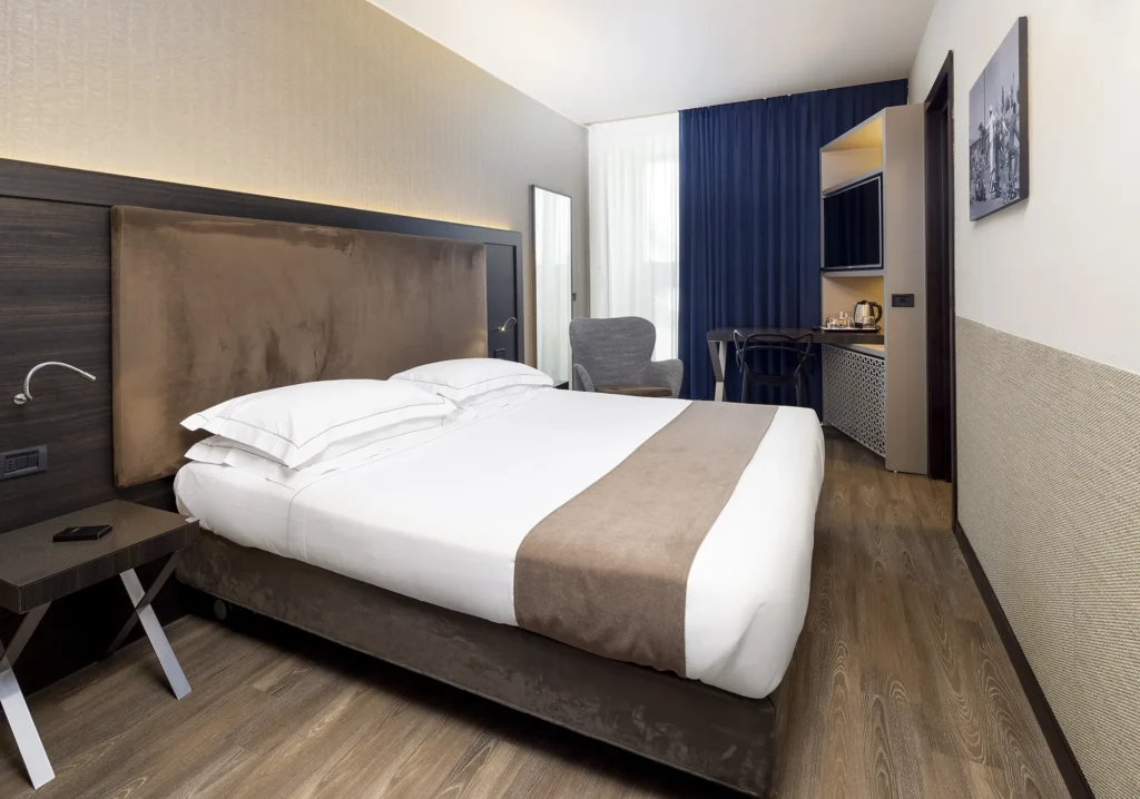 Nell'immagine, la camera Standard dell'Hotel Farnese: si vede il letto matrimoniale con di fianco una poltroncina e uno specchio a figura intera. Nell'angolo, una scivania con una sedia euna TV.